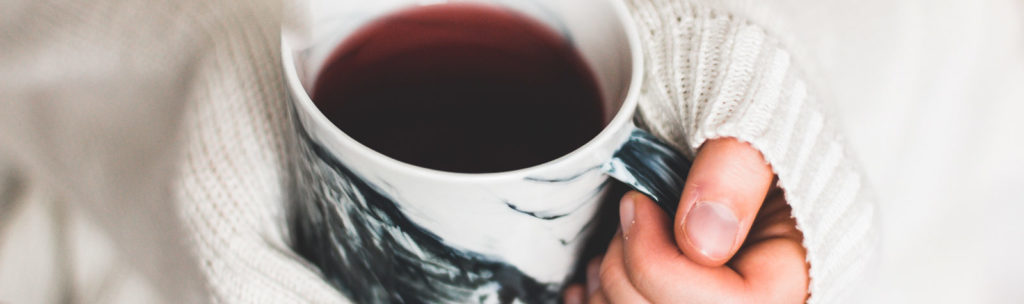 Hände einer Person, die eine schwarz-weiß marmorierte Tasse mit rotem Tee festhalten