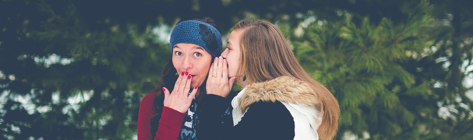 Eine junge Frau schaut gespeilt erschrocken und legt die Hand vor den Mund, während eine andere ihr etwas ins Ohr flüstert